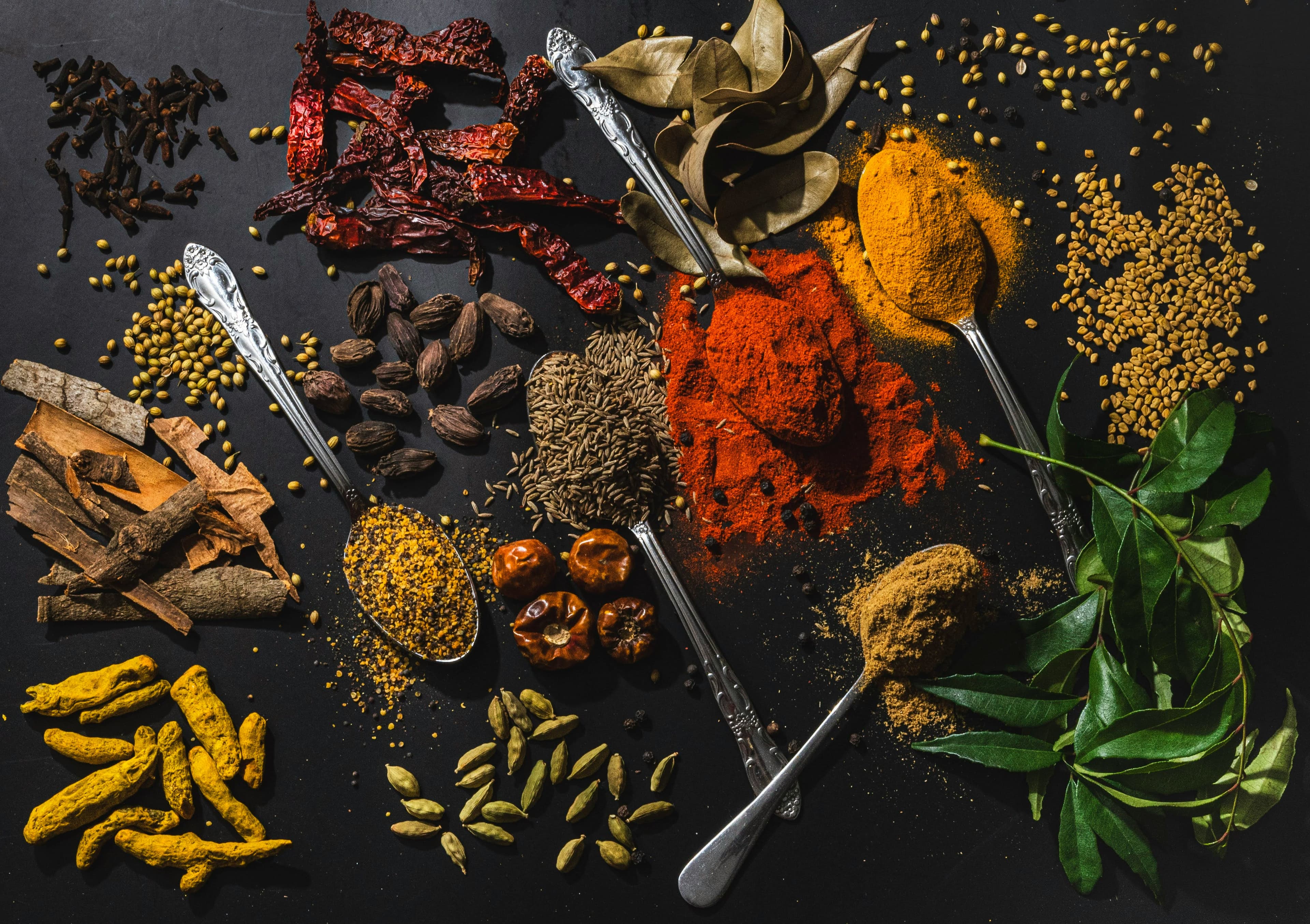 A vibrant flat-lay of heritage spices — turmeric, dried chillies, cardamom, cinnamon, cumin, fenugreek and fresh curry leaves on a dark surface